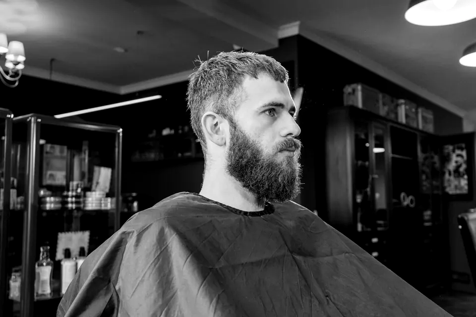 Schwarzweißfoto: Mann mit Bart sitzt im Friseurumhang vor einer Regalwand (Beispielbild)